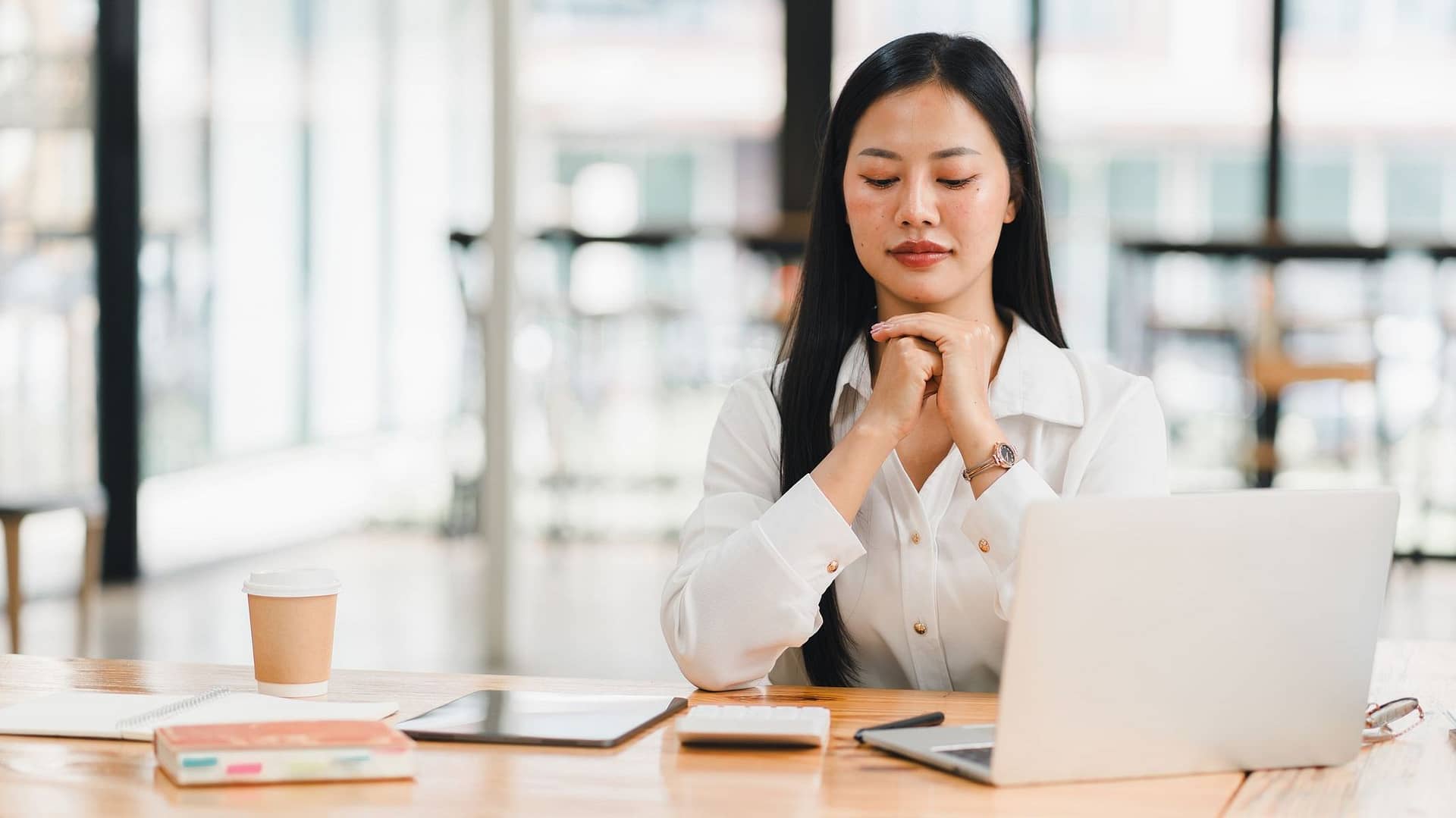 Focused woman in white shirt, sitting at desk with laptop, reflecting thoughtfully. Focused woman in white shirt, sitting at desk with laptop, reflecting thoughtfully